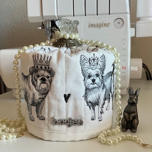 May include: White fabric drawstring bag featuring illustrations of a bulldog and a Yorkshire Terrier, both wearing crowns. The bag has a black heart and the word "boujee" in rhinestones. A pearl necklace and a small bronze rabbit figurine are also present.