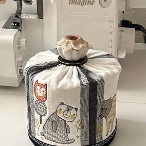 May include: A fabric spool cover with a whimsical forest animal design. The cover is white with black stripes and features embroidered bears and an owl. A decorative ceramic topper sits on top.