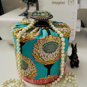 May include: A teal fabric drawstring bag with a floral pattern in yellow, pink, and black. A pearl necklace drapes over the bag, which is topped with a decorative knob. A small bronze rabbit figurine sits nearby.