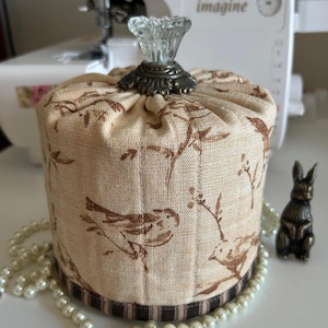May include: A cylindrical fabric container with a decorative glass knob on top. The beige fabric is patterned with brown birds and branches. A decorative border and a small bronze rabbit figurine are also present.