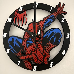 May include: A circular wall clock featuring a black frame with white numbers. The centre displays a red and blue graphic of a superhero in a crouching pose with a red spider emblem positioned near the eleven o'clock marker and black clock hands.
