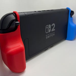 May include: A black Nintendo Switch console featuring a matte finish with the Nintendo Switch logo printed on the back. The device is equipped with a bright red protective grip on the left side and a vibrant blue protective grip on the right side.