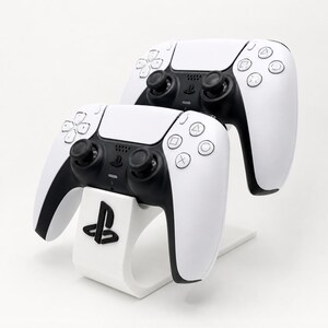 May include: Two white and black wireless game controllers positioned on a white curved display stand. The stand features a black PlayStation logo on the front. The controllers have dual analogue sticks, a directional pad, and four action buttons.