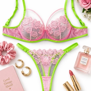 May include: A matching lingerie set featuring a pink lace bra and thong with bright lime green straps and trim. The bra has underwire cups with pink floral embroidery and thin pink accent straps. The thong features matching pink lace and green trim.
