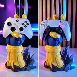 May include: A blue and yellow gloved hand sculpture with three metallic claws extending upward, designed as a controller stand. The base features jagged gray rock-like elements. The stand is shown holding a white Xbox controller and a white PlayStation controller.