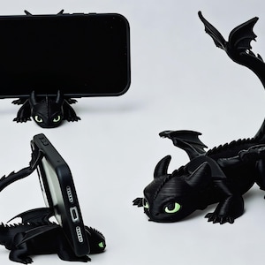 以下が含まれることがあります： 表面に質感のある黒いドラゴン型のスマートフォンスタンド。尖った耳と光る緑色の目が特徴です。長い曲がった尾と翼のデザインで、モバイル端末を横向きと縦向きの両方で支えることができます。