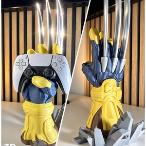 May include: A blue and yellow sculpted hand stand featuring three long silver metallic claws extending upward. The hand is designed to hold a white and black game controller. The base is decorated with gray jagged rock-like elements.