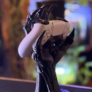 Può includere: Un supporto nero testurizzato a forma di mano artigliata che sostiene un controller per videogiochi wireless bianco. Il supporto presenta dita lunghe e appuntite e una robusta base circolare per una presa sicura.