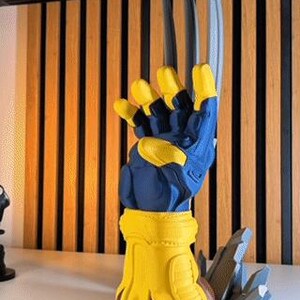 May include: A three-dimensional display piece featuring a blue and yellow glove with three long, silver-colored claws extending upward. The glove is mounted on a yellow base with brown accents, accompanied by gray, jagged, rock-like debris at the bottom.