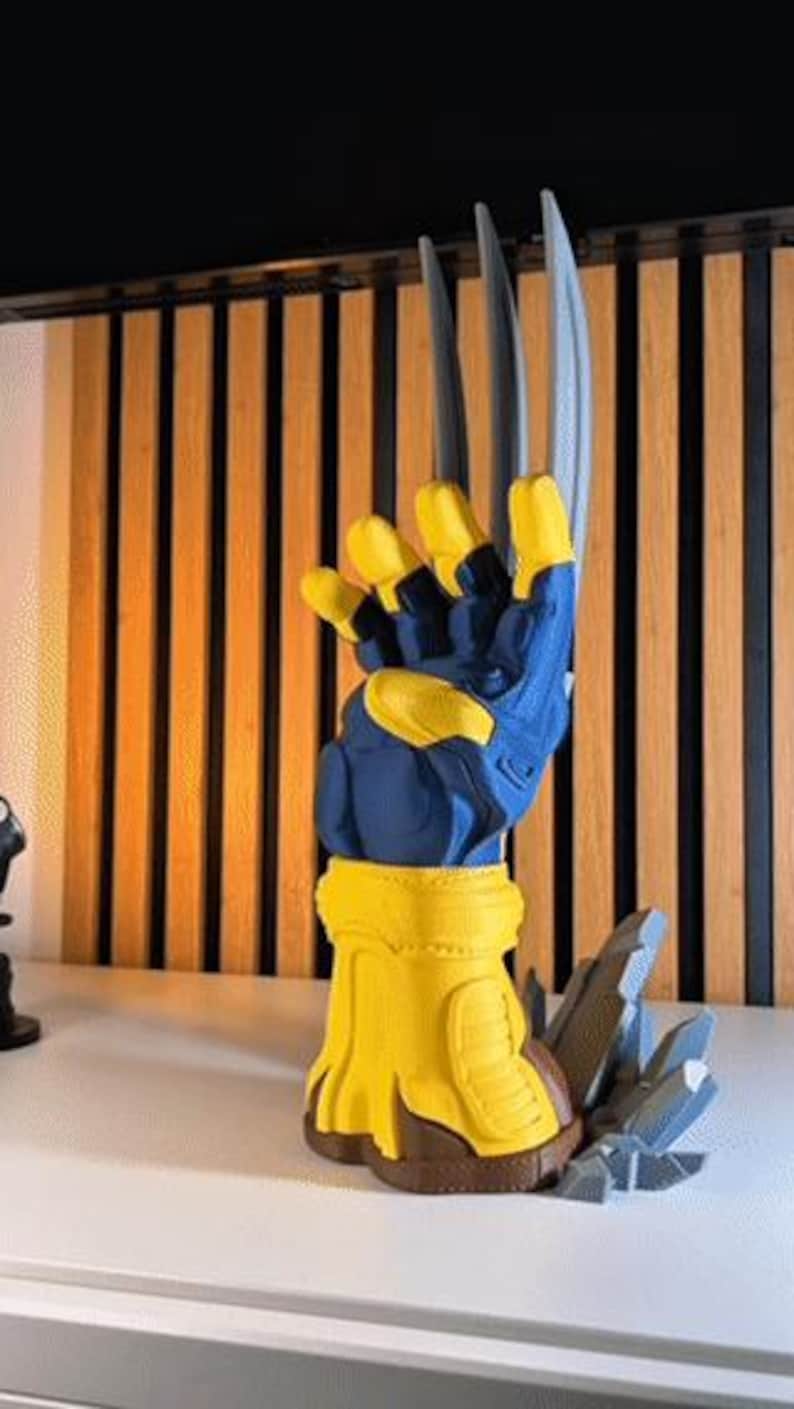 May include: A three-dimensional display piece featuring a blue and yellow glove with three long, silver-colored claws extending upward. The glove is mounted on a yellow base with brown accents, accompanied by gray, jagged, rock-like debris at the bottom.
