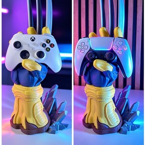 May include: A blue and yellow sculpted hand stand featuring three metallic claws extending upward, designed to hold a white Xbox controller or a white and black PlayStation controller. The base is shaped like jagged rock formations.
