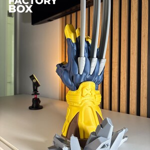 May include: A 3D printed display model of a blue and yellow glove featuring three long, silver-colored metallic claws extending from the knuckles. The glove rests on a jagged, grey rock-like base and includes an embossed X symbol on the wrist cuff.