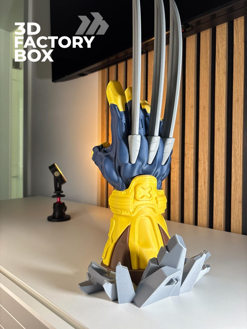 May include: A 3D printed display model of a blue and yellow glove featuring three long, silver-colored metallic claws extending from the knuckles. The glove rests on a jagged, grey rock-like base and includes an embossed X symbol on the wrist cuff.
