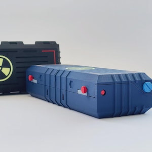 May include: A rectangular blue storage case with a yellow radiation symbol on top, featuring small red circular accents and a blue circular dial on the side, positioned in front of a black case with a matching yellow radiation symbol.