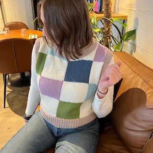 Op de afbeelding: Een mouwloos gebreid spencer vest met een groot ruitpatroon in wit, lichtblauw, lavendel en olijfgroen. Het kledingstuk heeft een geribbelde ronde hals en een geribbelde zoom in een neutrale beigetint, gedragen over een wit shirt met lange mouwen.