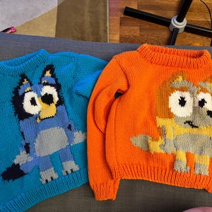 May include: Two long-sleeved knit sweaters. The blue sweater features a dark blue, light blue, and yellow cartoon dog character. The orange sweater features a yellow, tan, and white cartoon dog character. Both have ribbed necklines and cuffs.