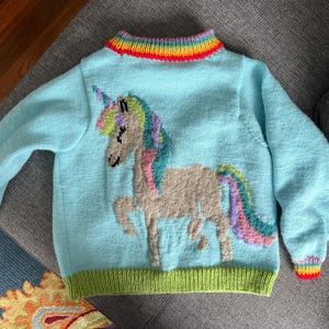 Op de afbeelding: Een lichtblauwe trui met lange mouwen en een gebreide eenhoornafbeelding met een veelkleurige manen en staart. De trui heeft een geribbelde kraag en boorden met een regenboogpatroon en een effen limoengroene geribbelde zoom aan de onderkant.