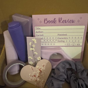 May include: A collection of stationery items in a box, including a purple book review card with fields for title and author, a floral bookmark, a heart-shaped floral tag, a purple page holder, a roll of purple tape, and a grey fabric hair scrunchie.
