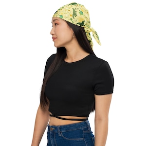Puede incluir: Una mujer con un pañuelo amarillo con un estampado floral y hojas verdes. El pañuelo está atado en la parte posterior de su cabeza. Lleva un top corto negro y pantalones vaqueros azules.