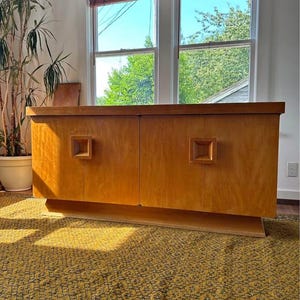 Può includere: Una credenza rettangolare in legno con finitura marrone chiaro, superficie superiore piatta e due ante. Ogni anta è dotata di una maniglia quadrata in legno incassata al centro del pannello. L'unità poggia su una base solida e rientrante.