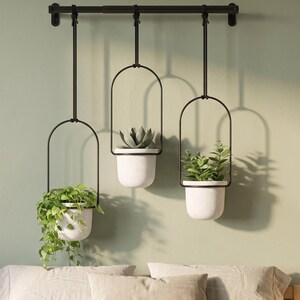 May include: A wall-mounted black metal rod with three hanging planters suspended by black cords. Each planter features a matte white speckled pot held within a black arched frame. The pots contain a trailing green plant, a succulent, and a leafy plant.