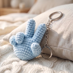 May include: A light blue crocheted hand gesture keychain featuring a peace sign shape. The soft, textured yarn figure is attached to a standard silver-toned metal split ring by a short chain, creating a compact accessory for keys or bags.