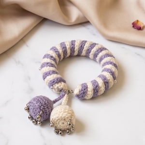 May include: A circular crochet accessory featuring alternating lavender and cream stripes. Two dangling crochet spheres, one lavender and one cream, are attached to the ring, each adorned with small silver-toned metal bells at the base.
