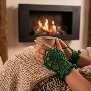 Può includere: Un paio di guanti senza dita in filato verde brillante, caratterizzati da un dettagliato motivo a ragnatela all'uncinetto che copre il dorso delle mani e si estende fino ai polsi con un bordo arricciato, mentre si tiene una tazza in ceramica chiara.