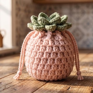 May include: A crochet drawstring pouch in a soft pastel pink color, featuring a textured stitch pattern and a decorative top element shaped like a green succulent plant with layered leaves. The pouch includes two matching pink yarn drawstrings.