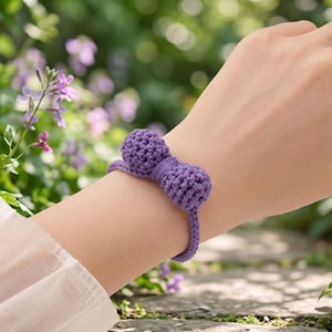 May include: A purple crochet bracelet featuring a central bow design made of two rounded, textured spheres connected by a thin, braided band. The accessory is crafted from yarn with a visible knit pattern, showcasing a consistent violet hue throughout.