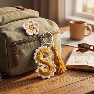 Puede incluir: Un llavero de ganchillo con una flor blanca de centro amarillo, una letra S de color amarillo mostaza con borde festoneado blanco y una borla a juego, todo unido a una anilla de metal plateado colgada de una mochila de color verde oliva.