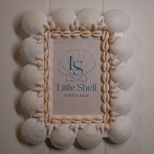 May include: A rectangular picture frame decorated with white scallop shells and smaller tan cowrie shells. The centre insert features a blue graphic of a shell with the letters LS and the text Little Shell COTTAGE printed in blue on a white background.