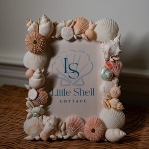 May include: A rectangular picture frame decorated with an assortment of natural seashells, including white, pink, and orange varieties. The centre of the frame displays a light-coloured insert with the blue text Little Shell COTTAGE and an LS logo.