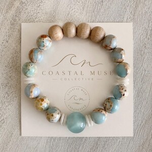 May include: A beaded bracelet featuring round light blue and brown mottled stone beads, four smooth light brown wooden beads, and small white disc spacers. The bracelet is displayed on a square card printed with the text Coastal Muse Collective.
