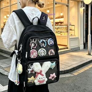 May include: A black backpack featuring two clear front pockets. The top pocket displays six circular anime character pins. The bottom pocket contains a plush white rabbit with a red checkered bow and four small plush stars in pink, blue, and yellow.