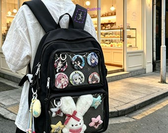 May include: A black backpack featuring two clear front pockets. The top pocket displays six circular anime character pins. The bottom pocket contains a plush white rabbit with a red checkered bow and four small plush stars in pink, blue, and yellow.