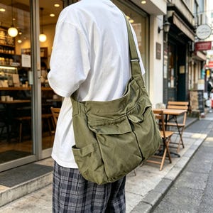 May include: An olive green canvas crossbody bag featuring a wide shoulder strap, two front pockets with flap closures, and side pockets. The bag is worn over a white t-shirt and plaid pants, showcasing a functional design with multiple storage compartments.