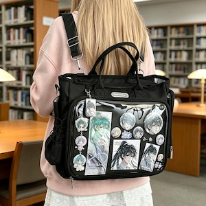 May include: A black ita bag backpack featuring a clear front display window filled with various anime-style character pins, rectangular cards, and hanging charms. The bag includes top handles, adjustable shoulder straps, and side pockets.