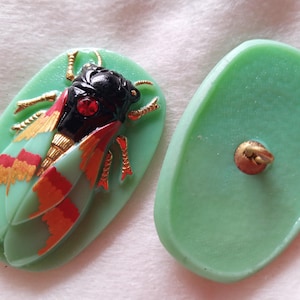 May include: Two-piece set of vintage green buttons. One button features a detailed insect design with black, red, and gold accents. The other button is plain green with a gold-coloured shank.