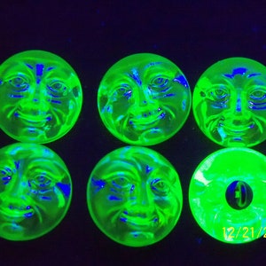 Peut inclure: Six boutons en verre vert avec un visage sculpté. Les boutons brillent sous une lumière noire, ce qui fait apparaître les visages en bleu. La date 12/21/2023 est écrite sur le bouton en bas à droite.