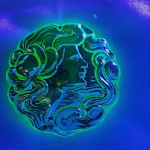 May include: A green glass cameo featuring a woman's face in profile. The cameo is set against a blue background.