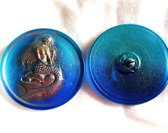 May include: Two round, blue buttons. One button features a mermaid design in silver and gold tones. The other button is a solid blue-green color with a shank on the back. These buttons are likely for sewing or crafts.
