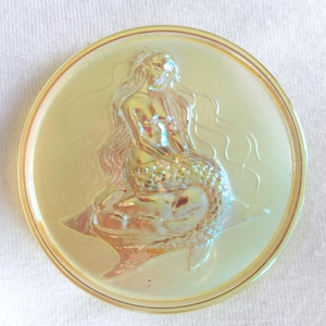 May include: A circular, iridescent plate featuring a raised relief of a mermaid. The mermaid is depicted seated, with flowing hair and a fishtail. The plate has a golden-yellow colour and a glossy finish, suitable for decorative purposes.
