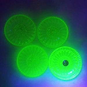 May include: Four circular glass buttons glowing with a bright neon green hue under ultraviolet light. Each button features a radial sunburst pattern radiating from the centre, with a small dark circular indentation in the middle of each piece.