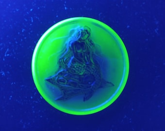 May include: A green glass paperweight with a mermaid design. The paperweight is round and has a smooth surface.