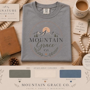 May include: A slate blue t-shirt featuring a graphic of mountains, a cross, and the text Mountain Grace Co. The shirt is displayed with a brown notebook, a coffee mug, a wooden bead garland, and a color chart showing bay, ivory, and blue jean options.