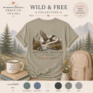 May include: A sage green t-shirt featuring a graphic of a mallard duck in flight over a mountain stream, with the text "MOUNTAIN GRACE CO. OUTDOORS". The shirt is displayed alongside a white mug with "faith grace adventure" and a beige backpack.