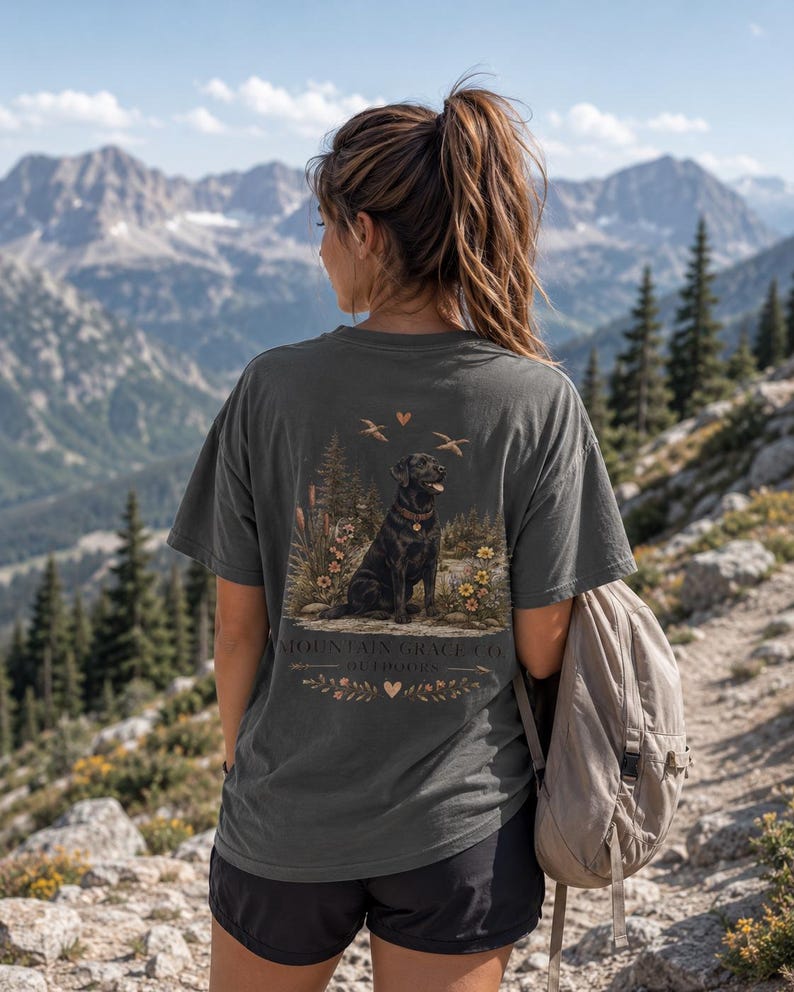 May include: A charcoal gray short-sleeve t-shirt featuring a graphic print on the back of a black Labrador retriever sitting in a forest landscape with flowers, birds, and the text Mountain Grace Co. Outdoors with small heart icons.