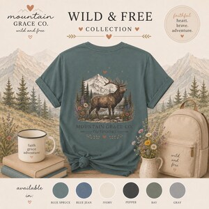 May include: A blue spruce t-shirt featuring a graphic of an elk in a mountain landscape with the text Mountain Grace Co. Outdoors. The shirt is displayed with a white mug labeled faith grace adventure and a beige backpack labeled wild and free.