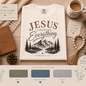 May include: A cream-colored t-shirt featuring the brown text Jesus Changes Everything and a mountain landscape graphic with pine trees and a lake, including the text Romans 8:28. Below are color swatches labeled Bay, Blue Jean, and Gray.